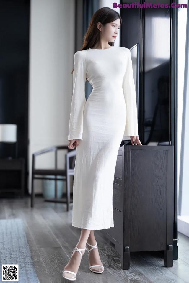 A woman in a white dress standing next to a dresser.