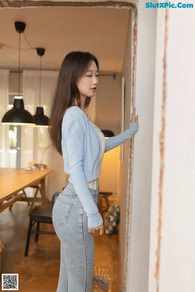 A woman in a blue sweater and jeans leaning against a wall.